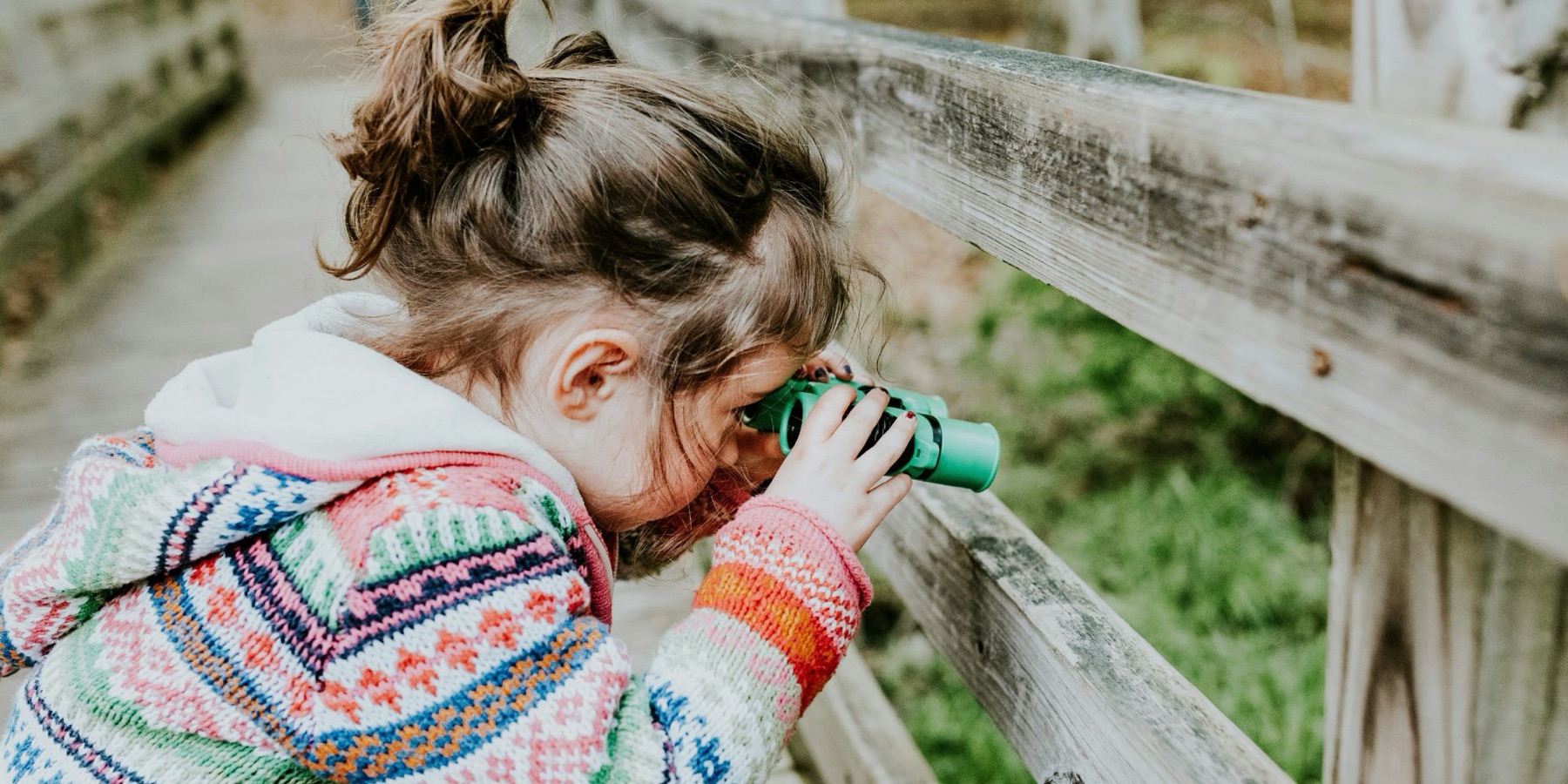 A little girl in a colorful sweater looking through green toy binoculars on a wooden bridge