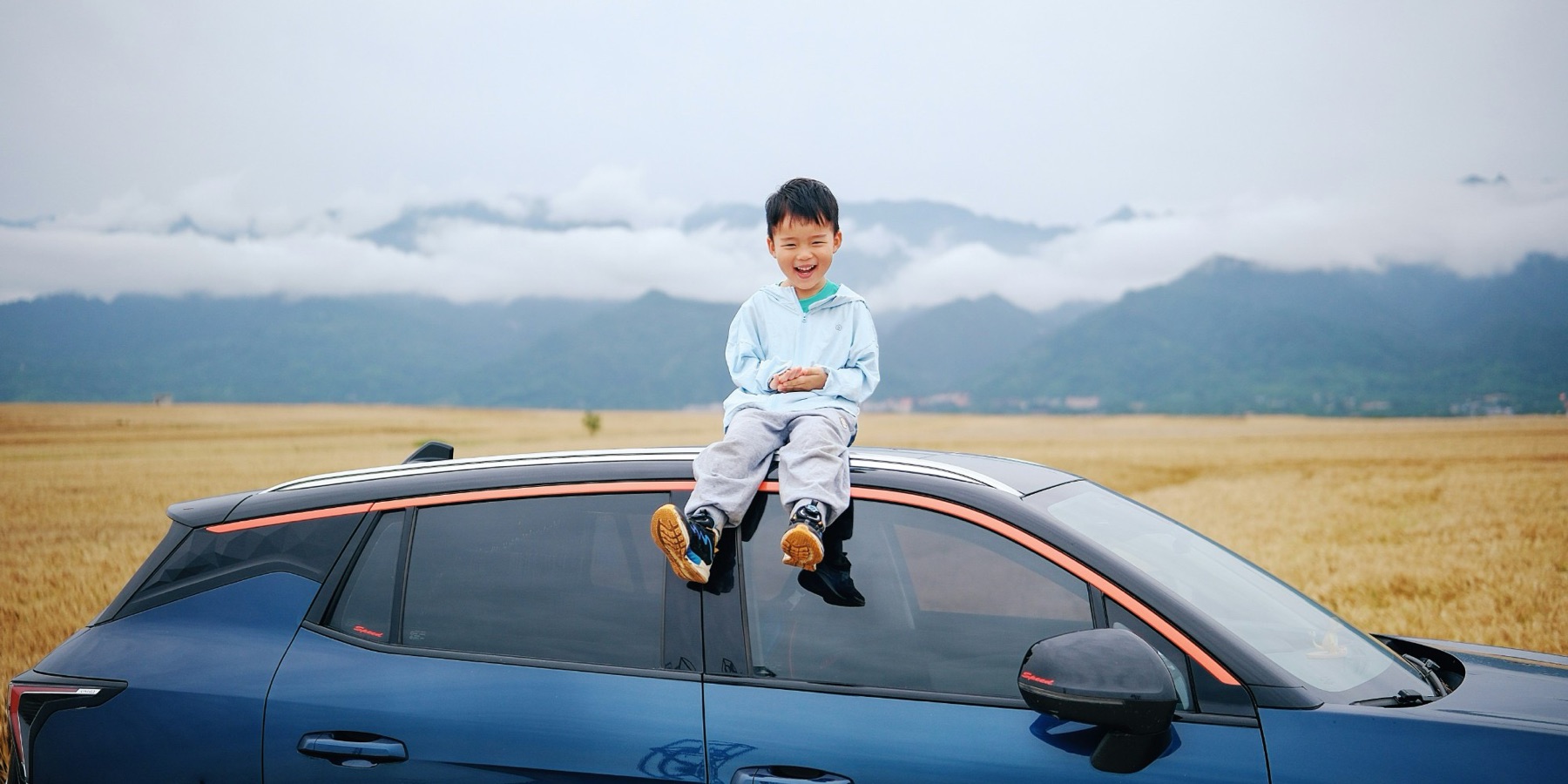 A laughing boy sitting on the roof of a car in front of golden fields and mountains