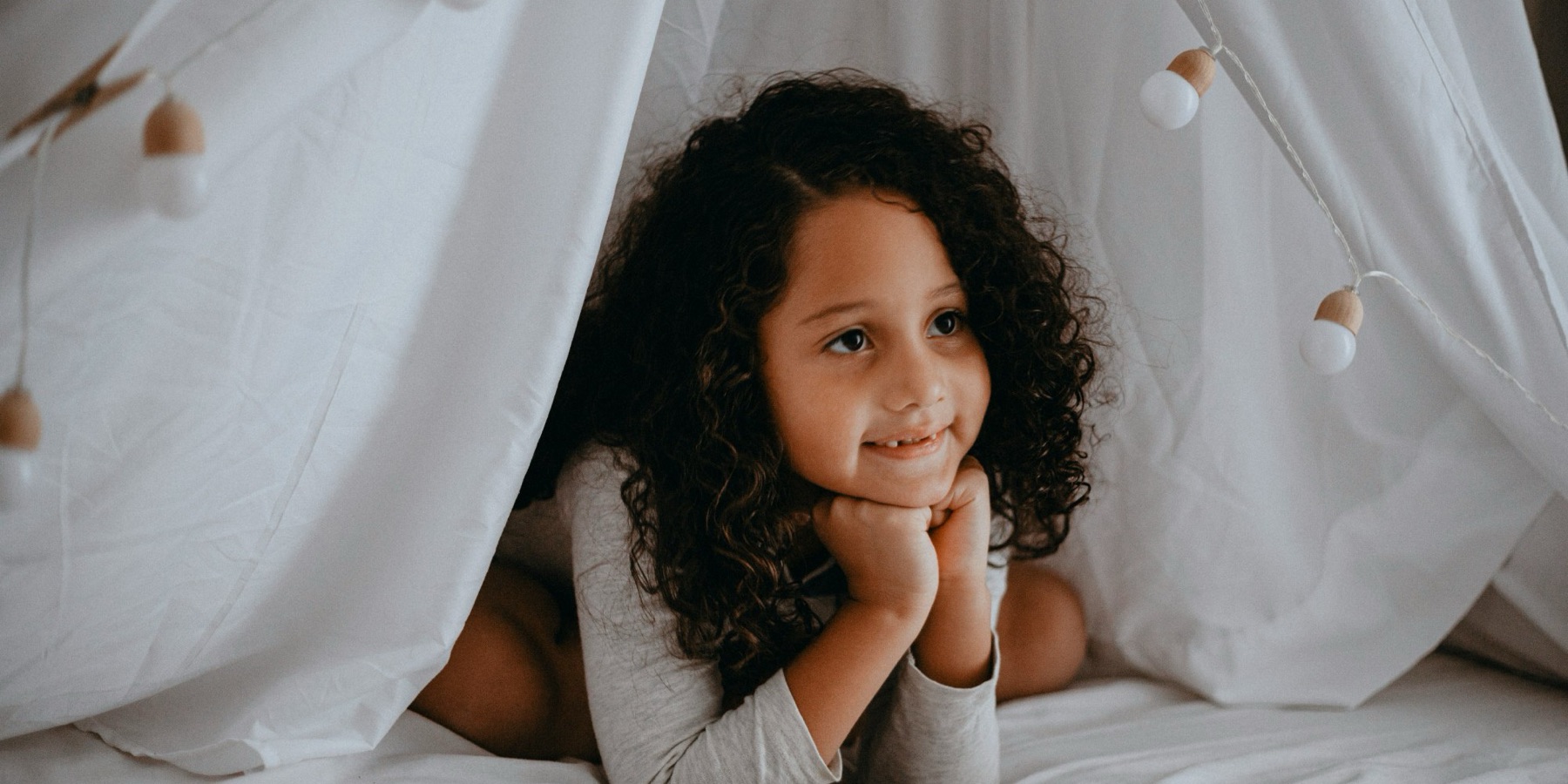 A smiling girl with curly hair lying in a cozy white play tent strung with lights